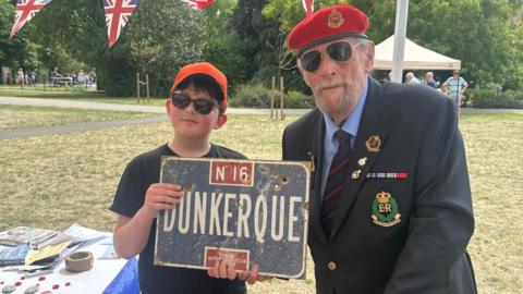 Ben, standing next to a Royal British Legion man, who is wearing a jacket and red beret. Ben has on glasses and an orange cap and is holding a sign. They are in a field, with grass all around them. 