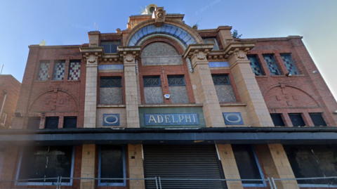 A large red brick building with stone accents, shutters covering doors and windows, and a security fence