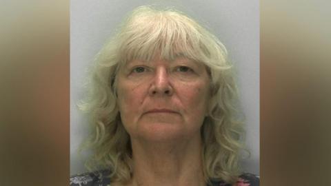 A mugshot of a woman in her 60s with wavy grey blonde hair and a fringe. She is straight faced as she stands in front of a pale grey background. From her shoulders, you can see she is wearing a floral T-shirt.