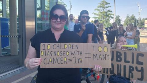 A woman is holding a cardboard banner saying too many children are being diagnosed with cancer. Another woman holds one calling for a public inquiry. Other people are gathered in the background.