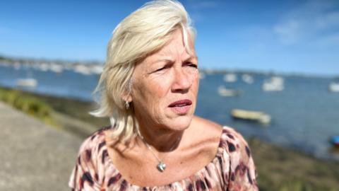 Melanie Leahy stands in front of Maldon harbour in Essex looking at the distance. She is wearing a pink and burgundy top with a silver heart locket around her neck. It is a sunny day. She has blonde, shoulder length hair. There are boats moored in the sea.
