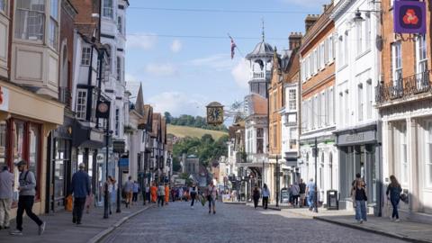 People walking on either side of a road in Guildford town centre. There is shops and businesses and blue skies. 