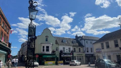 A general view of Fakenham town centre. Historic houses hold shops and cafes.
An ornate, black, Victorian-style lamp post is in the foreground. A few cars are parked on the road. People walk by.