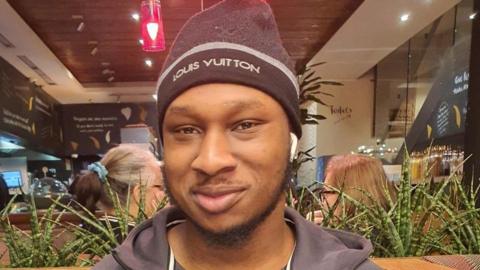 Khaleed Oladipo wearing a black hat and zip-up jacket, smiling. 