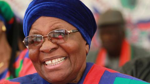 Netumbo Nandi-Ndaitwah smiles past the camera, wearing gold-rimmed glasses and the red, blue and green party colours of Swapo.