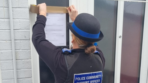 A police officer is seen sticking a piece of paper to the front door of a property.