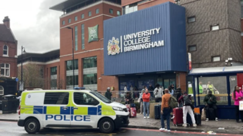 A police van parked outside a college building with a big blue sign saying 'University College Birmingham'. There are around a dozen people outside the building which also has a bus shelter in front of it.
