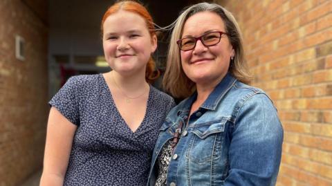 A teenage girl with red hair stands next to her Mum who wears a denim jacket
