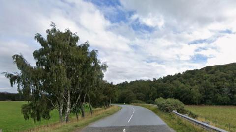Picture of the B851, with trees on either side of the road and a blue sky with white clouds.