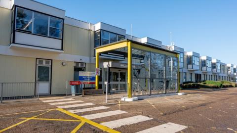 External view of the Queen Elizabeth Hospital in King's Lynn. There is a pedestrian crossing on the road in front of the entrance which is covered by a glazed canopy with yellow supports. 