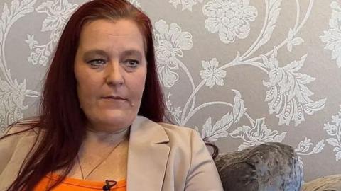 Rebecca Ramsay with long brown hair wearing a biege blazer and orange top sitting on a velvet chair with floral patterned wallpaper in the background.