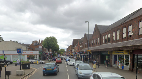 A high street busy with cars, with a road junction to the left. An acorns shop can be seen to the left, with a range of shop fronts on the right. Buildings on both sides are modern two to three storey structures built with dark red/brown brick. Pedestrians can be seen on both sides of the pavement.