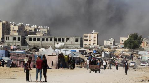 Smoke rises following an Israeli strike on a residential building in Khan Younis in Gaza. There are tents set up in the foreground and people walking around.