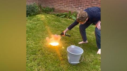 A still showing a teenage boy bending over with a tool as a small explosion is pictured on the ground. He is standing on the grass in a back garden, with a bucket next to the fire.