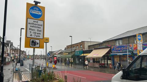 A view of the traffic-restricted zone on Portswood Road in Southampton. A red block of colour is seen on the road indicating the start point of the restricted zone and a sign detailing the restrictions is on the left hand side. Shops are visible on the opposite side of the road.