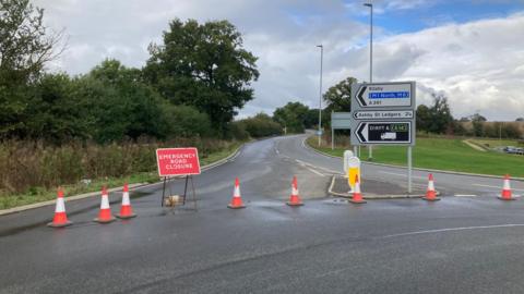 Exit from roundabout coned off, with red "emergency road closure" sign. There is also a roadsign to Kilsby, Ashby St Ledgers and DIRFT. There are trees on one side of the road and grass on the other.