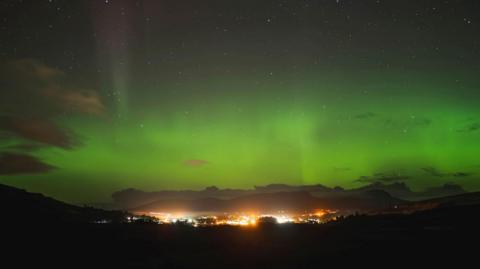 The aurora, also known as the Northern Lights, glows green above a brightly lit community in the Isle of Skye.