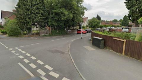 A Google street view image of the entrance to a road from a T-junction. It is a residential street with houses and fences on either side, with a large tree to the left. Cars are parked along the side of the road in the distance.