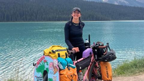 Naomi Hancocks stands in front of blue water with her pink bike which is laden with all the equipment she needs to travel solo.