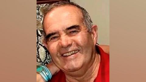 A smiling man with short grey hair and black eyebrows, wearing a red top 