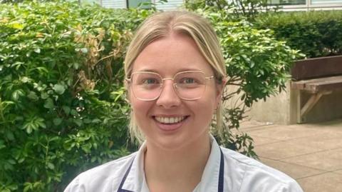 Becky Sheppard is sitting on a bench in front of a bush. She has blonde hair in a ponytail and is wearing a white hospital uniform. She has clear framed glasses.