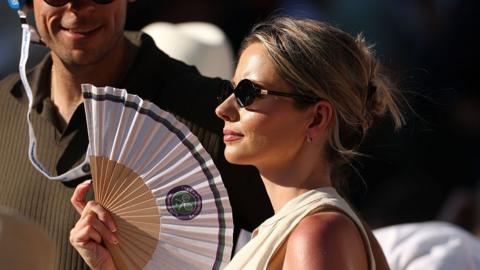 A woman wearing black sunglasses fans themselves at Wimbledon 2025.