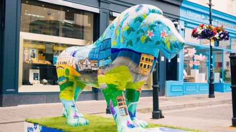 A large sculpture of a lion, painted with landmarks from across Gloucestershire, along with water and floral scenery, sits on a high street in a town centre.