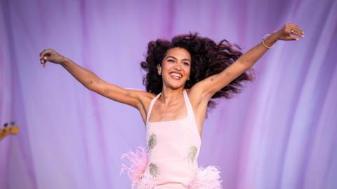 Olivia Dean stands with her arms wide, smiling. Her long dark hair is blowing to the side. She's wearing a pink dress.