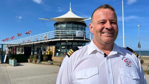 A man wearing a white shirt with naval-style epaulettes smiles as he stands in front of a modern-looking building with a glass tower, canopy and five Union flags flying under a blue sky with the sea in the background.   