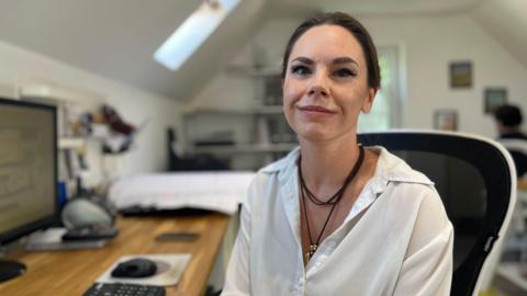 A woman with dark brown hair tied back sat at her desk looking into the camera. She is wearing a white button down shirt with a black string necklace with gold pendant. 