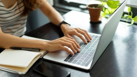 A pair of hands resting on a laptop's keyboard, with an empty notepad and pen and mobile phone on a table next to it