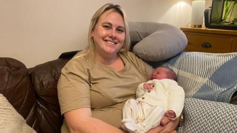 A blonde woman wearing a light-brown coloured t-shirt is smiling and cradling her new born baby in her arms. The baby is dressed in a white romper with giraffes on. They are sat on a sofa and there is a neck pillow propped up behind her.