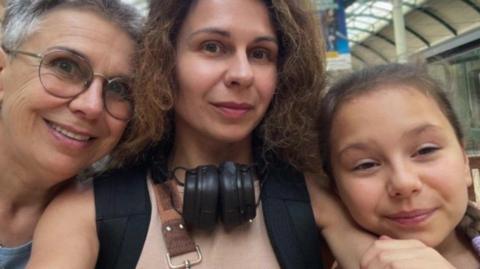 A woman is taking a selfie with a young girl to the left of her with her hand on her arm and an older woman to the right with short grey hair and glasses. They are all smiling and the background is blurred but appears to be a train station. The woman in the middle has long brown hair and is wearing black headphones around her neck.