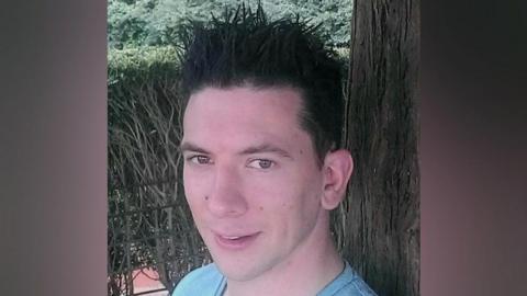 A man with black hair that is spiked up, and is wearing a light blue T-shirt, he is standing in front of a tree and a hedge