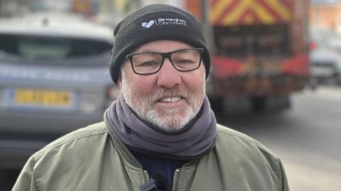 A middle-aged man wearing a black woolly hat with Birmingham City Council written on the front smiles into the camera. He is wearing black-rimmed glasses, a scarf and a green jacket. 