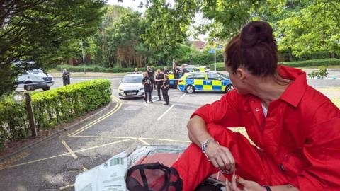 A Palestine Action protester sits atop a van wearing a bright red boiler suit, with a metal chain around their wrist. They are looking out on three police cars parked a short distance away, and a number of uniformed police officers. 