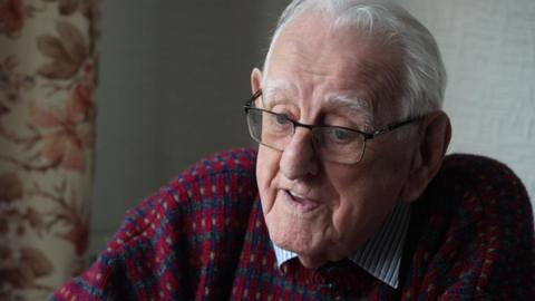 Tony Johnson wearing a blue and white shirt under a purple jumper talks at his home to the BBC