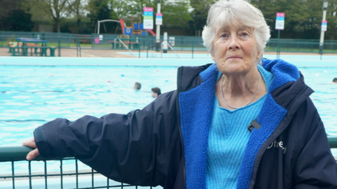 Pat Hemsley, who is wearing a navy blue coat and a blue jumper, standing by the poolside. She is smiling at camera. 