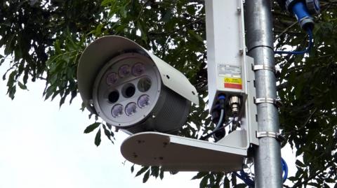 We can see a circular modern camera with multiple lenses on it. It's installed on a pole, with a tree visible behind it.