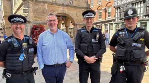 Four men, three of them in black police uniform and the fourth wearing a blue shirt and blue trousers, standing in a town square with a stone building standing on pillars behind them