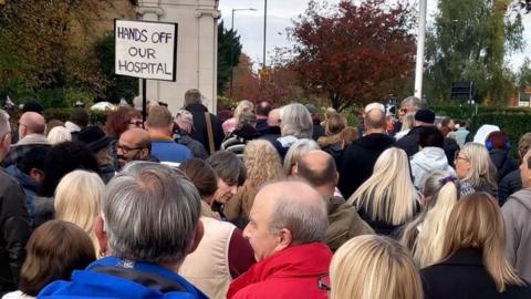 A crowd of dozens of people gather in front a large park area. A sign with "Hands Off Our Hospital" is visible in the background.