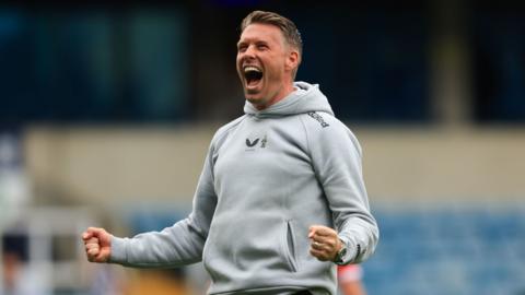 Rob Edwards has mouth wide open smiling and holds both hands in fists in celebration. he wears a grey hoodie.