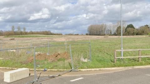 A road is at the bottom of the image which has a silver fence on the left blocking off parts of the road, and a wooden fence to the right. Behind them is a field which has trees on the other side in the background. 