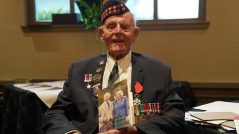 Veteran Jim Glennie in uniform with medals, smiling and saluting. He is holding a card featuring an image of the King and Queen on the cover