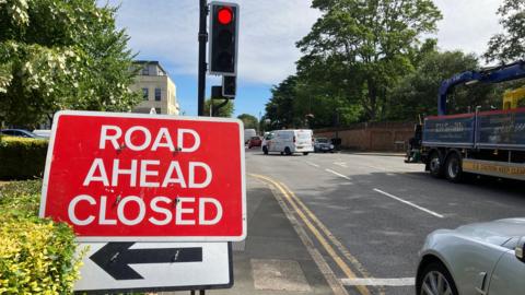 Red traffic sign indicating "Road ahead closed" at the side of a road in front of a red traffic light. A pavement hedge is on the left and a silver car is parked at the junction on the right. Vehicles can be seen crossing the junction.