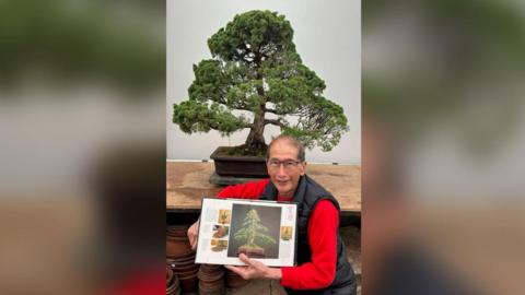 Peter Chan (an older man with glasses smiling at the camera) stands in front of a bonsai tree while holding a poster with a bonsai tree on