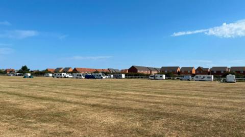 More than 10 white caravans have camped on a field of dry grass in front of a housing estate in Redcar. The caravans are spread out along the far side of the field and the sky is clear and blue.
