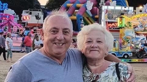 A man and a woman standing arm in arm in front of a fairground