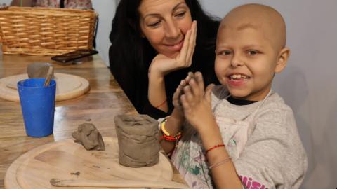 Girl with clay on her hands with a pottery structure on a table. Her mother, to her right, looks lovingly at her