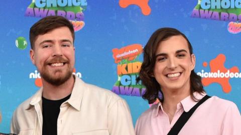 MrBeast pictured with Ava Kris Tyson at the Nickelodeon Kids Choice Awards. MrBeast has short brown hair and a short beard and wears a white overshirt over black T-shirt. Ava has shoulder-length brown hair parted to the side and wears a pink shirt. 